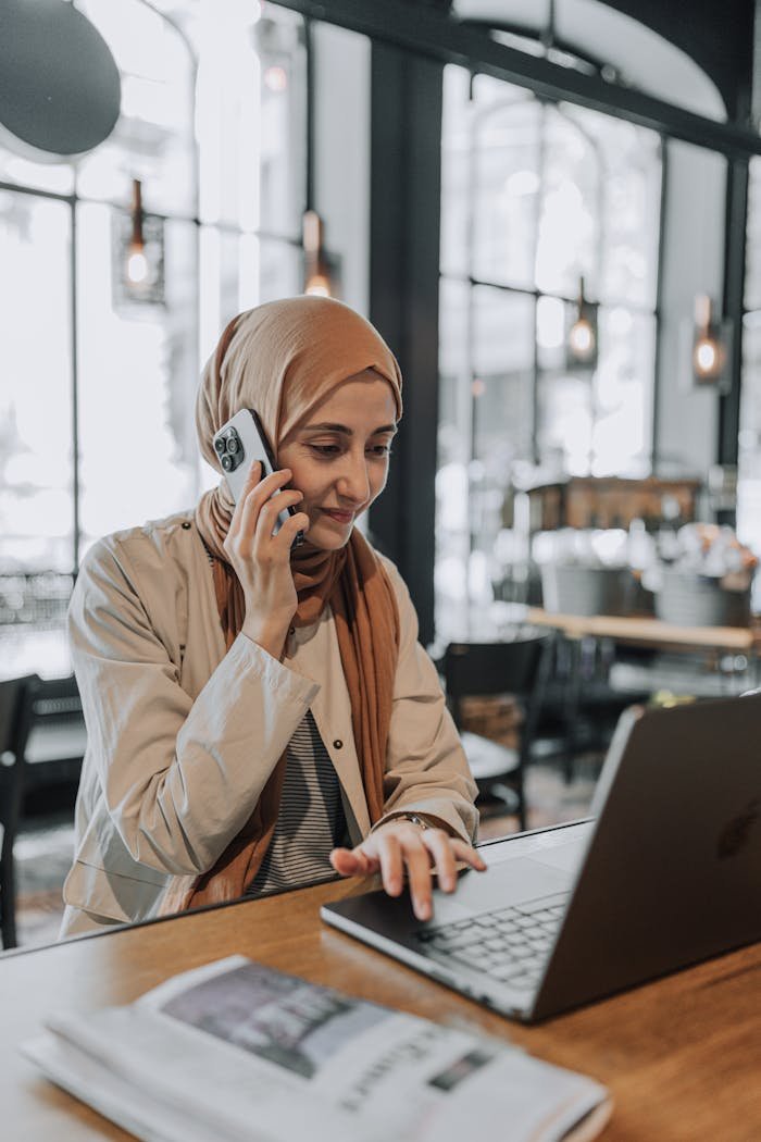contact-img A woman in a hijab multitasks with a phone and laptop in a stylish café, showcasing modern working culture.