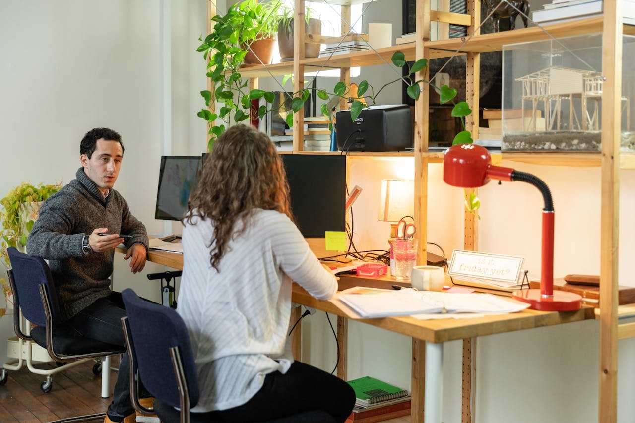 services-01 Two colleagues discuss work at a modern office desk with multiple monitors and greenery.